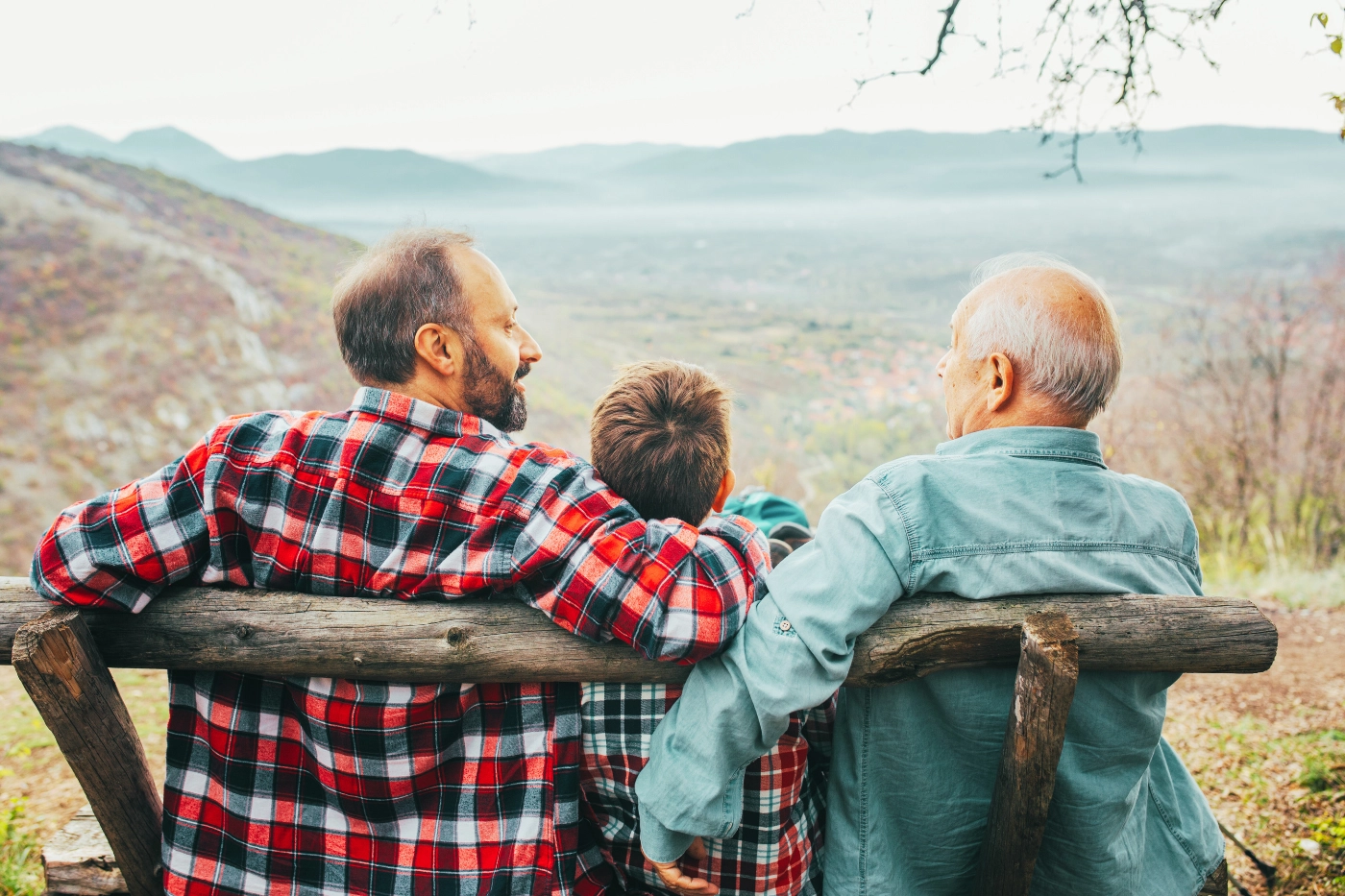 A grandfather sits with his son and grandson, overlooking a scenic mountain view while discussing transferring their assets into a trust.