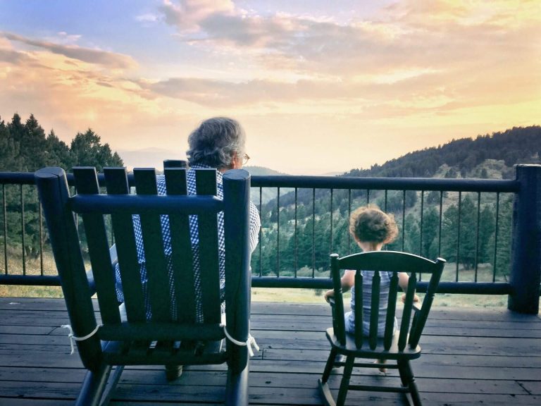 An elderly man and a young child sit in rocking chairs on a deck, looking out at a scenic mountain sunset, showcasing moments of distinctive care services.