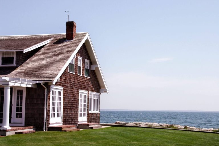 A traditional Cape Cod style coastal house with brown cedar shingle siding and white-trimmed windows, situated on a green lawn next to the blue ocean under a clear sky.
